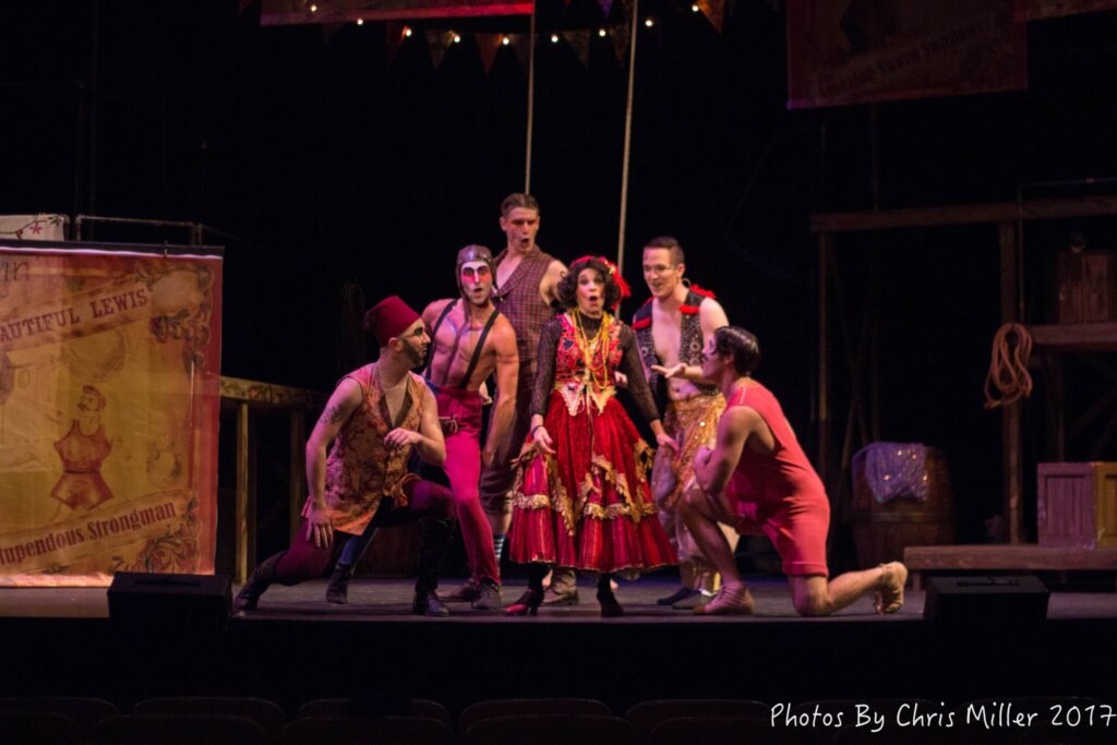 Darrell Wood performs centerstage in a circus-themed production, surrounded by dancers in vibrant red and gold costumes under theatrical lighting.