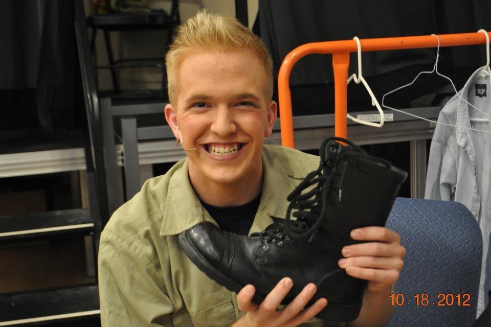 Darrell Wood, grinning, proudly holds a large black combat boot while seated backstage, with costume racks and gear in the background.