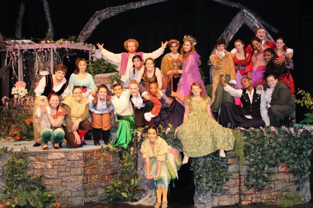 Cast members in fairytale costumes pose on an outdoor stone set, with Darrell Wood among them dressed as a whimsical character in a forest-themed production.