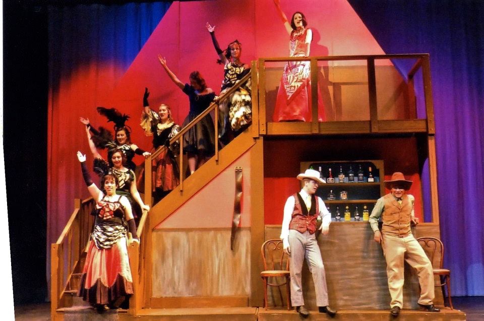 A lively western-themed stage performance in mid-action, featuring Darrell Wood and ensemble on a multi-level set with bright red and gold lighting.

