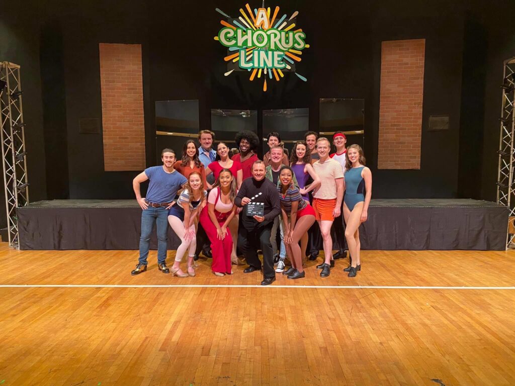 The cast of “A Chorus Line,” including Darrell Wood, pose on a dance studio stage in full costume in front of a glowing show logo backdrop.