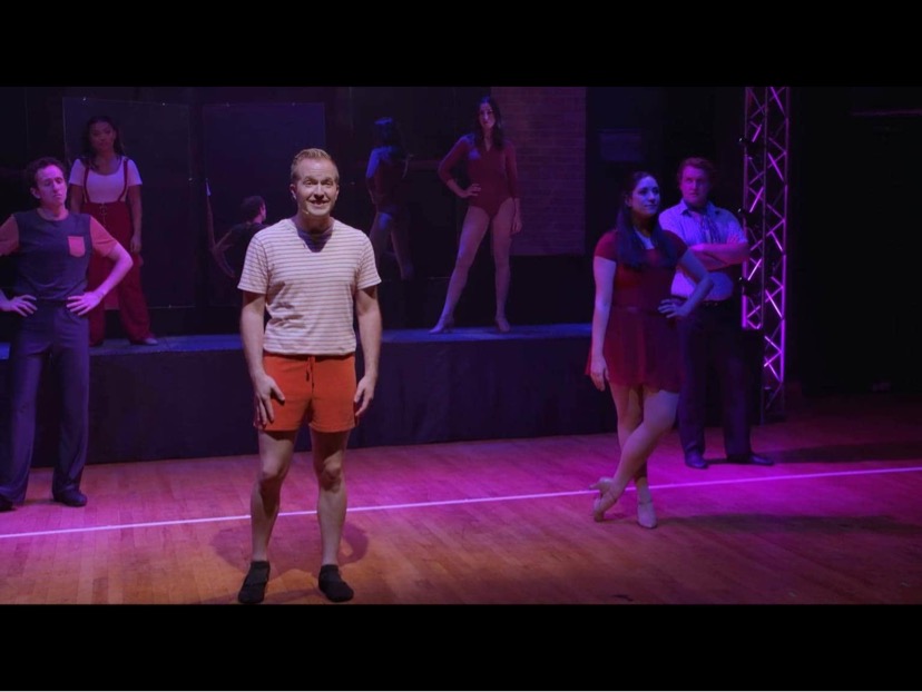 Darrell Wood performs confidently centerstage in rehearsal attire—striped shirt and red shorts—lit by dramatic pink and purple stage lighting during a musical number.