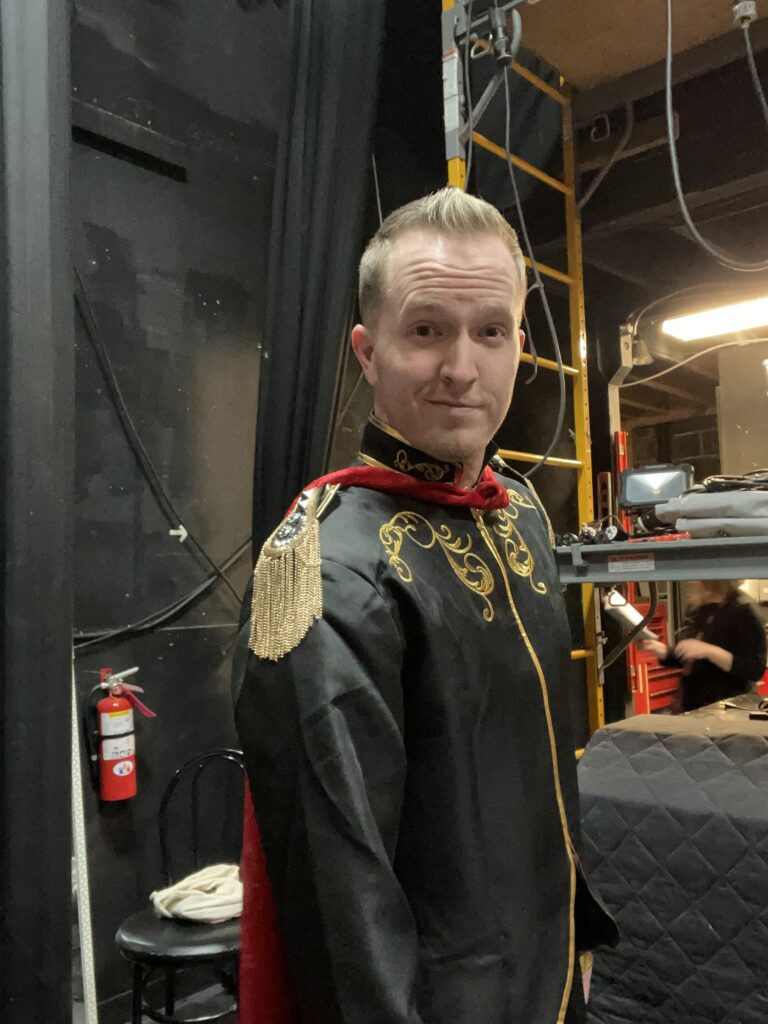 Darrell Wood stands backstage in an elaborate black and gold military-inspired costume, preparing for his next scene.