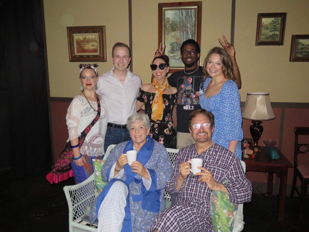 Darrell Wood poses with a diverse cast in pajamas and loungewear in a cozy backstage lounge, all smiling and holding mugs in a celebratory group photo.


